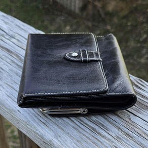 May include: Black leather wallet with a silver clasp and a snap closure. The wallet has white stitching around the edges and a flap with a silver snap. The wallet is sitting on a wooden surface.