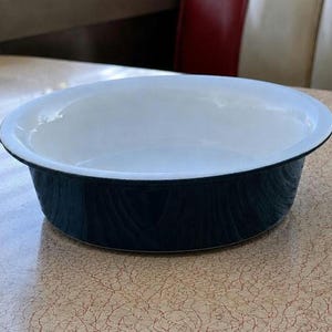 May include: A round, white-interior bowl with a dark blue exterior. The bowl has a wide rim and sits on a light-colored surface. The bowl is likely made of enamel or a similar material. The design is simple and classic.