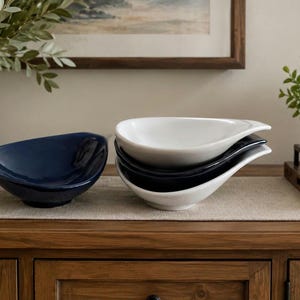 May include: A collection of ceramic serving bowls in navy blue, black, and white. The bowls have a unique teardrop shape and are stacked on a wooden surface. Decorative elements include a vase with greenery and a small potted plant.