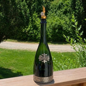 May include: A dark green glass bottle with a gold-colored pourer spout. The bottle features a silver-toned ornate crest emblem on the front and a silver-toned metal base band with the embossed text Segura Viudas.