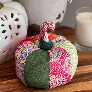 May include: A fabric pumpkin crafted from various colorful patterned patches, including floral, solid green, and gingham prints. The pumpkin features a stem with a red and white striped pattern, sitting alongside a white ceramic pumpkin and a candle.