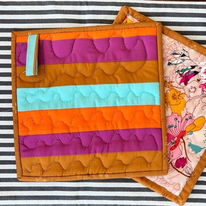 May include: Two square quilted pot holders. One has horizontal stripes of orange, teal, purple, and brown with a teal loop. The other has a floral pattern with pink, yellow, and white flowers on a light pink background, both with brown borders.