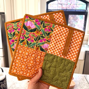 May include: A set of two square quilted pot holders featuring a patchwork design. The fabric patterns include a yellow background with pink and green floral motifs, an orange and white gingham check, a solid orange floral print, and a sage green leaf print.