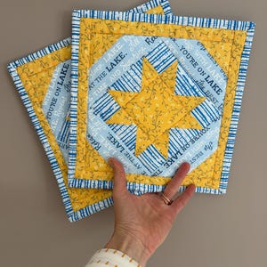 Op de afbeelding: Drie vierkante, gewatteerde pannenlappen met een gele ster in het midden. De pannenlappen hebben een blauw-wit gestreepte rand en een geel bloemenpatroon. De tekst op de pannenlappen luidt "You're on Lake".