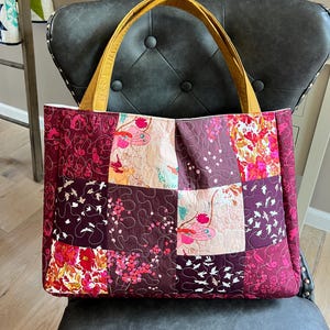 May include: A quilted tote bag with a patchwork design in shades of burgundy, pink, and cream. The bag features floral and bird patterns, with mustard-yellow handles. The bag is sitting on a dark gray chair.
