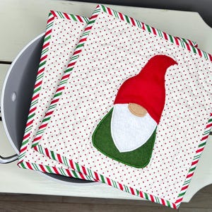 May include: Two square quilted pot holders with a Christmas gnome design. The pot holders have a white background with red polka dots and a red, green, and white striped border. The gnome has a red hat, white beard, and green body.