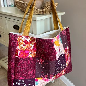 Peut inclure: Un sac fourre-tout patchwork avec un motif matelass&eacute;. Le sac pr&eacute;sente un m&eacute;lange de motifs floraux et abstraits dans des tons bordeaux, roses et oranges. Il a des anses dor&eacute;es et un int&eacute;rieur blanc. Le sac mesure environ 40 cm de large et 36 cm de haut.