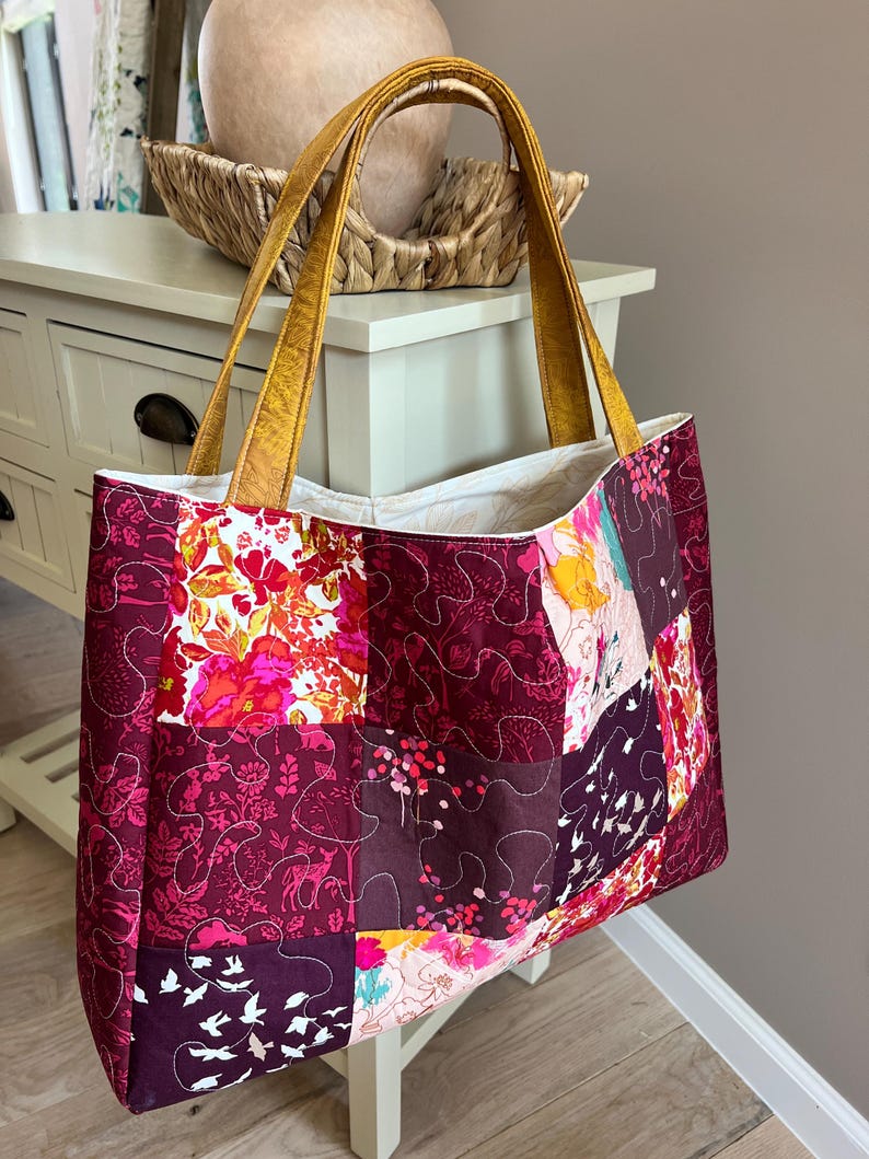 Peut inclure: Un sac fourre-tout patchwork avec un motif matelass&eacute;. Le sac pr&eacute;sente un m&eacute;lange de motifs floraux et abstraits dans des tons bordeaux, roses et oranges. Il a des anses dor&eacute;es et un int&eacute;rieur blanc. Le sac mesure environ 40 cm de large et 36 cm de haut.