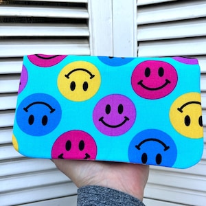 May include: A rectangular clutch with a vibrant turquoise background, covered in colorful smiley faces. The smiley faces are in shades of yellow, pink, purple, and blue, each with a black smile and eyes. The clutch is held in a hand.