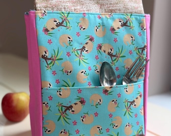May include: A rectangular fabric utensil holder featuring a light blue background with a repeating pattern of brown sloths and pink flowers. The pouch has bright pink side panels, a beige patterned top flap, and a front pocket containing a metal spoon and fork.