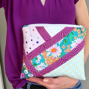 Puede incluir: Un colorido estuche con cremallera de patchwork con un estampado floral y de lunares. El estuche tiene una cremallera blanca y se sostiene contra una camisa morada. El diseño incluye telas rosas, turquesas, moradas y blancas.