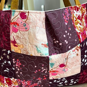 Peut inclure: Un sac fourre-tout patchwork avec un motif floral dans les tons de rose, bordeaux et cr&egrave;me. Le sac pr&eacute;sente des carr&eacute;s matelass&eacute;s avec divers motifs floraux et un fond bordeaux uni. Les anses sont de couleur marron clair.