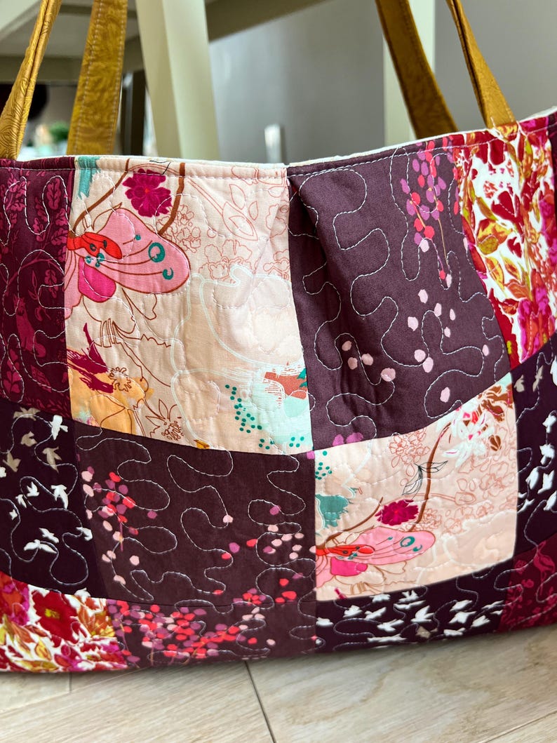 Peut inclure: Un sac fourre-tout patchwork avec un motif floral dans les tons de rose, bordeaux et cr&egrave;me. Le sac pr&eacute;sente des carr&eacute;s matelass&eacute;s avec divers motifs floraux et un fond bordeaux uni. Les anses sont de couleur marron clair.