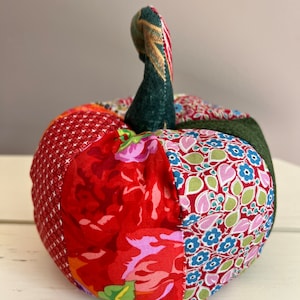 Può includere: Una zucca di patchwork con uno stelo verde. La zucca è realizzata con diversi tessuti con motivi floreali e geometrici in tonalità di rosso, rosa, blu e verde.