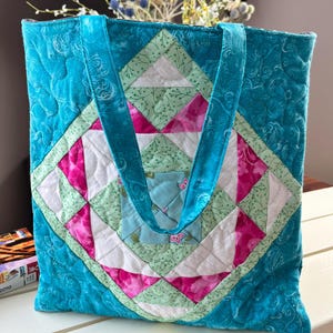 以下が含まれることがあります： ターコイズ色のキルティングトートバッグ。ピンク、グリーン、ホワイトの幾何学模様が施されています。バッグにはターコイズ色のストラップが2本付いています。