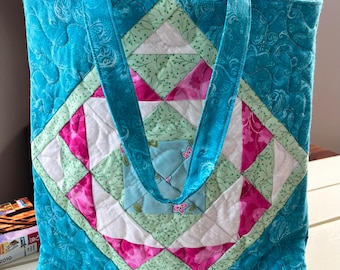 Puede incluir: Una bolsa de mano acolchada turquesa con un patrón geométrico en tonos de rosa, verde y blanco. La bolsa tiene dos asas turquesas.