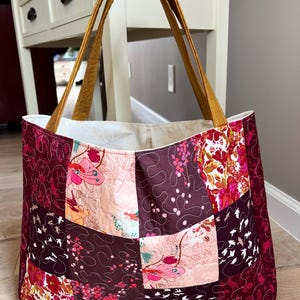 Peut inclure: Grand sac fourre-tout matelass&eacute; avec un motif patchwork. Le sac pr&eacute;sente des motifs floraux et abstraits dans les tons bordeaux, rose et cr&egrave;me. Il a des anses couleur moutarde et un int&eacute;rieur cr&egrave;me.