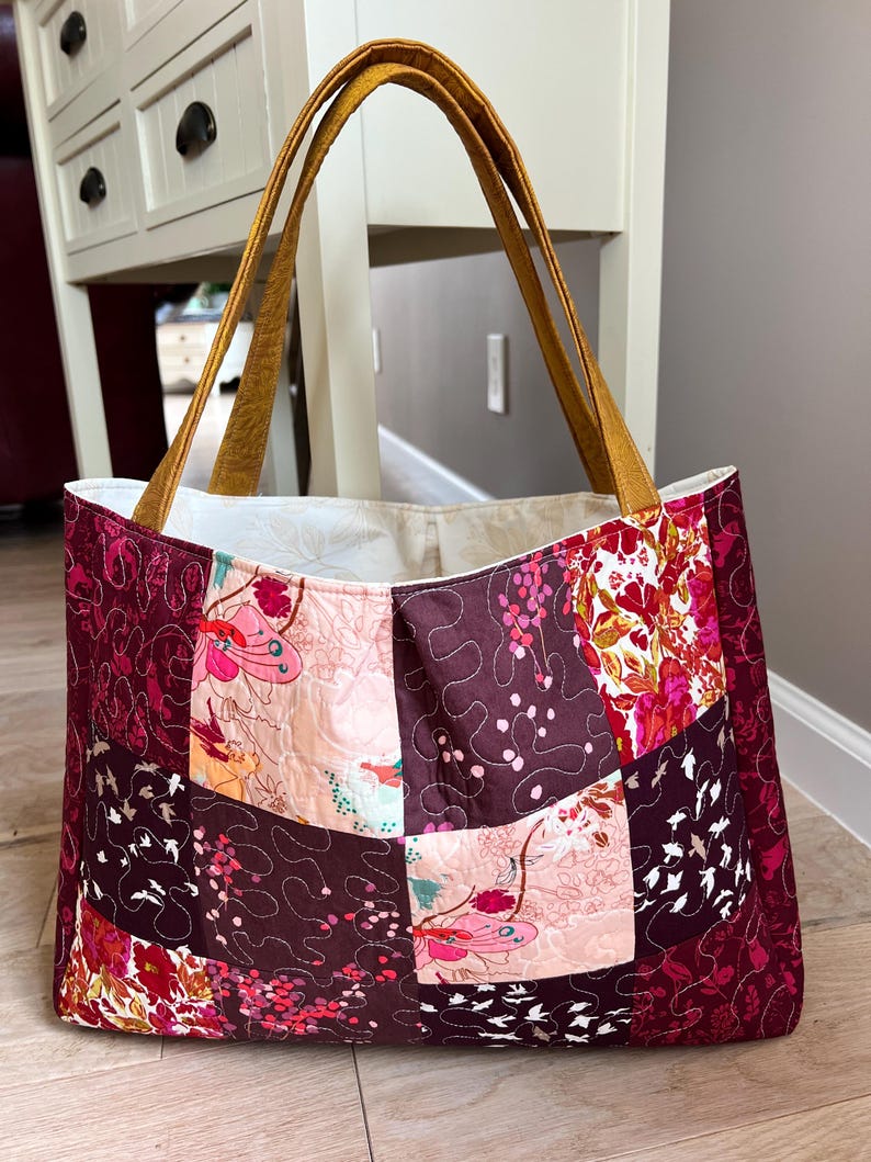 Peut inclure: Grand sac fourre-tout matelass&eacute; avec un motif patchwork. Le sac pr&eacute;sente des motifs floraux et abstraits dans les tons bordeaux, rose et cr&egrave;me. Il a des anses couleur moutarde et un int&eacute;rieur cr&egrave;me.