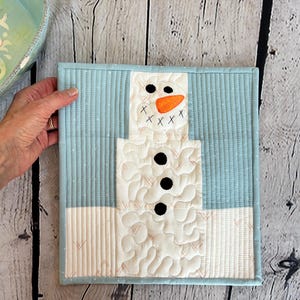 May include: A quilted snowman wall hanging. The snowman is made of white fabric with black button eyes, a carrot nose, and a stitched mouth. The background is light blue with vertical lines and a light blue border. The piece is square.