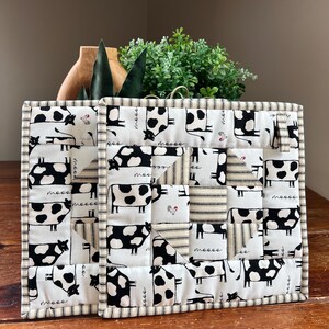 May include: A quilted hot pad featuring a patchwork design with black and white cow print fabric. The border is striped, and the fabric has the word "moo" printed on it. The hot pad is on a wooden surface.