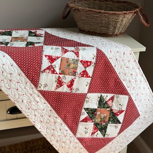 May include: A quilted table runner featuring a patchwork design with alternating star blocks and solid red dotted fabric panels. The quilt is stitched with a white floral pattern and includes blocks with dark green and red floral star motifs.