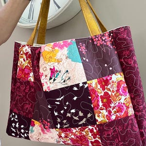 Peut inclure: Grand sac fourre-tout patchwork avec des carr&eacute;s de tissu &agrave; motifs floraux et d'oiseaux dans les tons bordeaux, rose, jaune et turquoise. Le sac a des anses en cuir couleur moutarde.