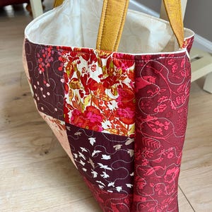 Peut inclure: Sac fourre-tout matelass&eacute; avec un motif patchwork. Le sac pr&eacute;sente des tissus dans les tons de rouge, bordeaux et rose, avec des motifs floraux et d'oiseaux. Les anses sont de couleur jaune moutarde.