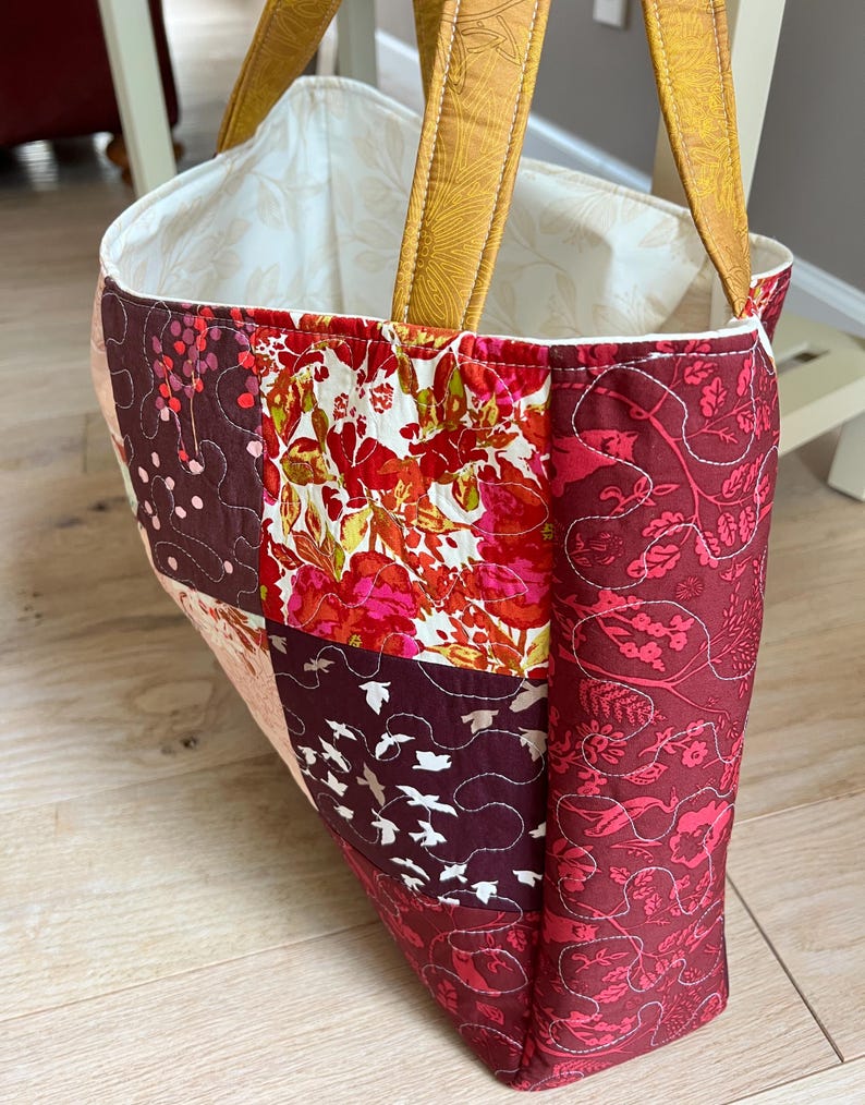 Peut inclure: Sac fourre-tout matelass&eacute; avec un motif patchwork. Le sac pr&eacute;sente des tissus dans les tons de rouge, bordeaux et rose, avec des motifs floraux et d'oiseaux. Les anses sont de couleur jaune moutarde.