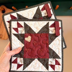 May include: Two square quilted potholders featuring a central dark red star pattern with a small cream heart appliqué. The design includes cream fabric with floral prints, dark brown accents, and a small fabric loop for hanging on each piece.
