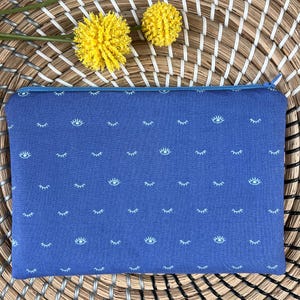 Könnte beinhalten: Eine blaue Reißverschlusstasche mit einem weißen Augen- und Wimpernmuster. Die Tasche liegt auf einer geflochtenen Strohoberfläche.