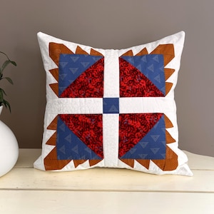 May include: A white pillow with a geometric pattern in red, blue, and brown. The pillow has a white background with a central square made of four smaller squares. Each smaller square is made of a different fabric, with a red and blue pattern in the corners and a brown fabric along the edges.