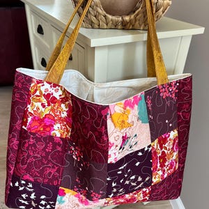 Peut inclure: Un sac fourre-tout patchwork avec un motif floral dans les tons bordeaux, rose et jaune. Le sac a des anses dor&eacute;es et un int&eacute;rieur cr&egrave;me. Le sac est fait de tissu matelass&eacute;.