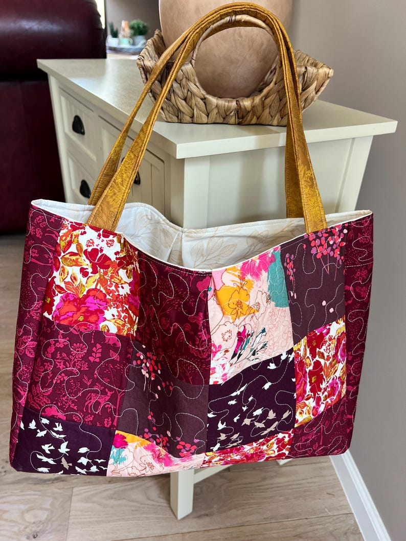 Peut inclure: Un sac fourre-tout patchwork avec un motif floral dans les tons bordeaux, rose et jaune. Le sac a des anses dor&eacute;es et un int&eacute;rieur cr&egrave;me. Le sac est fait de tissu matelass&eacute;.
