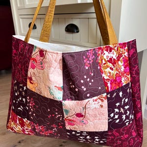 Peut inclure: Un sac fourre-tout patchwork avec un motif matelass&eacute;. Le sac pr&eacute;sente divers motifs floraux et abstraits dans des tons bordeaux, rose et cr&egrave;me. Il a des poign&eacute;es en cuir beige et un int&eacute;rieur blanc.