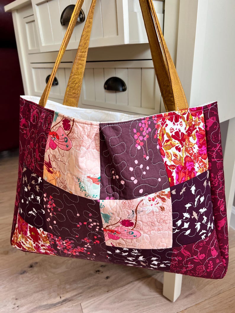 Peut inclure: Un sac fourre-tout patchwork avec un motif matelass&eacute;. Le sac pr&eacute;sente divers motifs floraux et abstraits dans des tons bordeaux, rose et cr&egrave;me. Il a des poign&eacute;es en cuir beige et un int&eacute;rieur blanc.