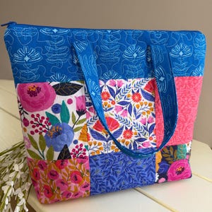 Op de afbeelding: Een gewatteerde tote bag met een levendig bloemen patchwork ontwerp. De tas heeft een blauwe rits en handvatten, met een mix van roze, blauwe en witte bloemenpatronen. De tas is ongeveer 35 cm breed en 30 cm hoog.