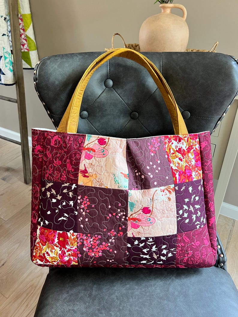 Peut inclure: Un sac fourre-tout matelass&eacute; avec un motif patchwork dans les tons bordeaux, rose et cr&egrave;me. Le sac pr&eacute;sente des motifs floraux et d'oiseaux, avec des anses jaune moutarde. Le sac est pos&eacute; sur une chaise gris fonc&eacute;.