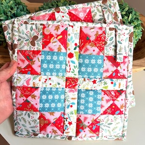 May include: A square quilted potholder featuring a patchwork design with red, pink, and blue fabric squares. The pattern includes gingerbread men, snowflakes, pinecones, and holly leaves. It has a white background with a fabric hanging loop.
