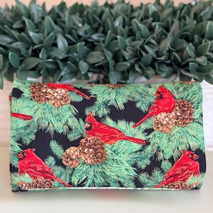 May include: A rectangular fabric clutch featuring a repeating pattern of bright red cardinal birds perched among green pine branches and brown pinecones against a solid black background. The accessory has a structured, folded design.