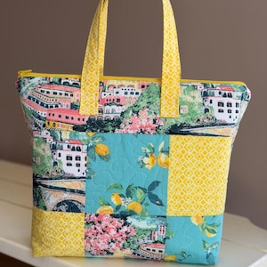Op de afbeelding: Een gewatteerde draagtas met een gele rits en handvatten. De tas heeft een patchwork-ontwerp met een kuststadscène, citroen- en bloempatronen en gele geometrische vierkanten. De tas staat op een wit oppervlak.