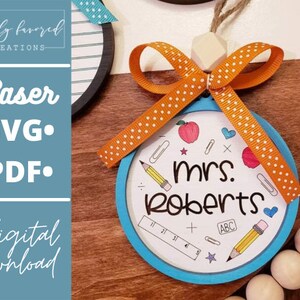 May include: A blue and white round ornament with the text "mrs. Roberts" and school-themed illustrations. The ornament is decorated with an orange ribbon with white polka dots.