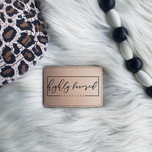 May include: A brown leather label with a stitched border and the text "highly favored creations" in black cursive font.
