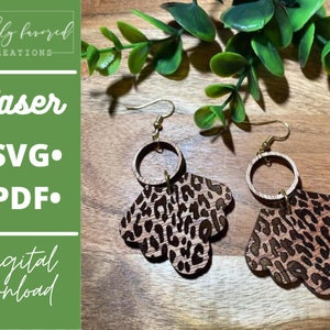 May include: A pair of laser cut wooden earrings with a leopard print design and gold hoops. The earrings are on a wooden surface with green foliage in the background. The text "Laser Cut SVG PDF" is visible in the image.