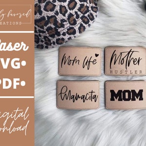 Pode incluir: Quatro patches retangulares de couro castanho claro com texto preto: "Mom life", "Mother Hustler", "Mamacita" e "MOM". Os patches estão sobre uma superfície branca e fofa. Um item com estampa de leopardo é visível no fundo.