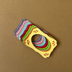 May include: A stack of colorful paper cutouts with a yellow frame and a central oval opening. The cutouts are layered, revealing a spectrum of colors including pink, blue, green, and red. The yellow frame has decorative cut-out details.