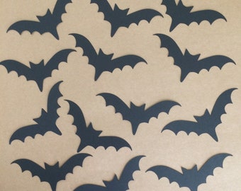 May include: A collection of 14 black paper bats, cut out in a simple silhouette shape, scattered on a brown background.