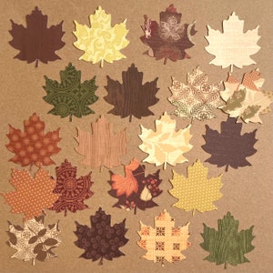 May include: A collection of twenty die-cut paper maple leaves in various patterns and colors, including shades of brown, cream, yellow, orange, and green. The leaves feature diverse designs such as polka dots, wood grain, floral, and geometric prints.
