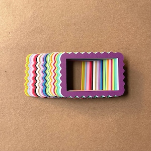 May include: A stack of colorful paper frames with scalloped edges. The frames are layered on top of each other, creating a rainbow effect. The top frame is purple and has a smaller, rectangular opening in the center.