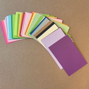 May include: A fan of colorful cardstock paper in various shades of blue, green, yellow, orange, pink, purple, brown, and black. The paper is arranged in a semi-circle with the top of the fan facing the viewer. The bottom of the fan is a solid purple piece of cardstock.