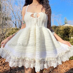 May include: A white crochet dress with a lace trim. The dress has a sweetheart neckline and a tie at the front. The dress is made with a textured crochet stitch.